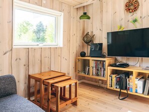 A cozy living room with a wooden floor, a large window, a flat-screen TV, a bookshelf, and a small wooden table.