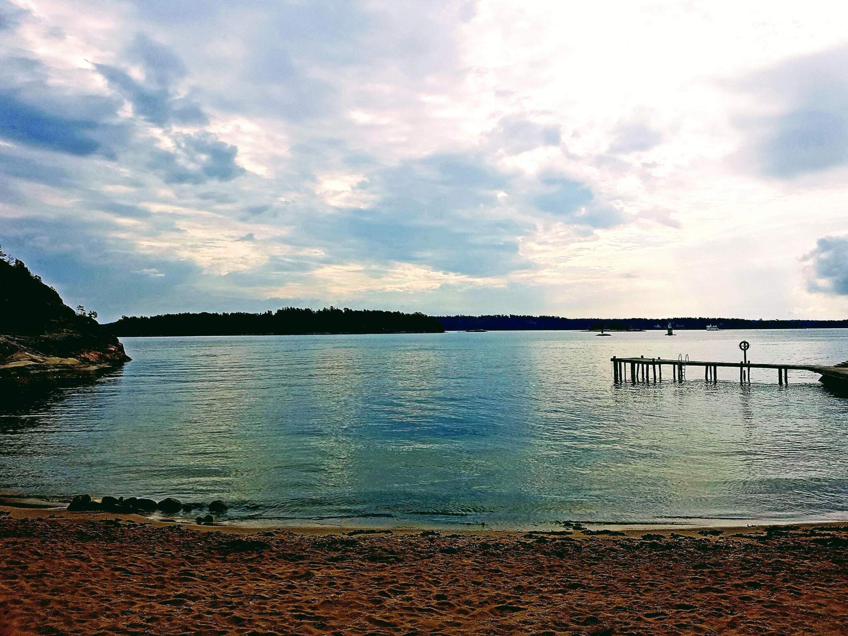 A calm lake with a wooden pier extending into the water, surrounded by a rocky shoreline and distant trees.