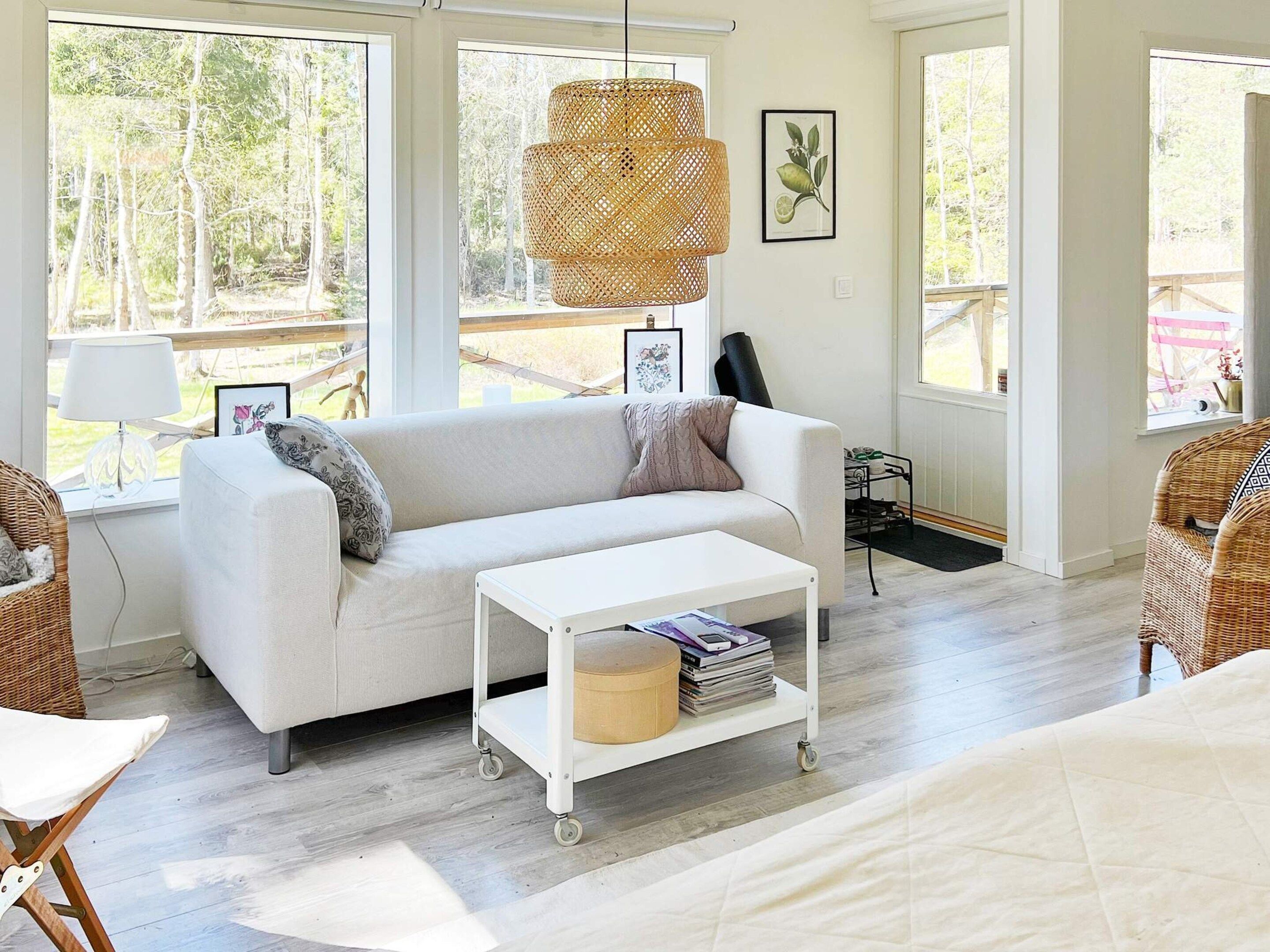 Un salón con sofá blanco, una mesa de centro blanca con revistas, sillas de mimbre y ventanas grandes con vista a árboles.