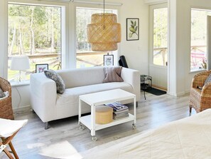 Un salón con sofá blanco, una mesa de centro blanca con revistas, sillas de mimbre y ventanas grandes con vista a árboles.
