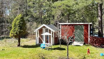 A small wooden cabin with a porch and a shed in a grassy area surrounded by trees.