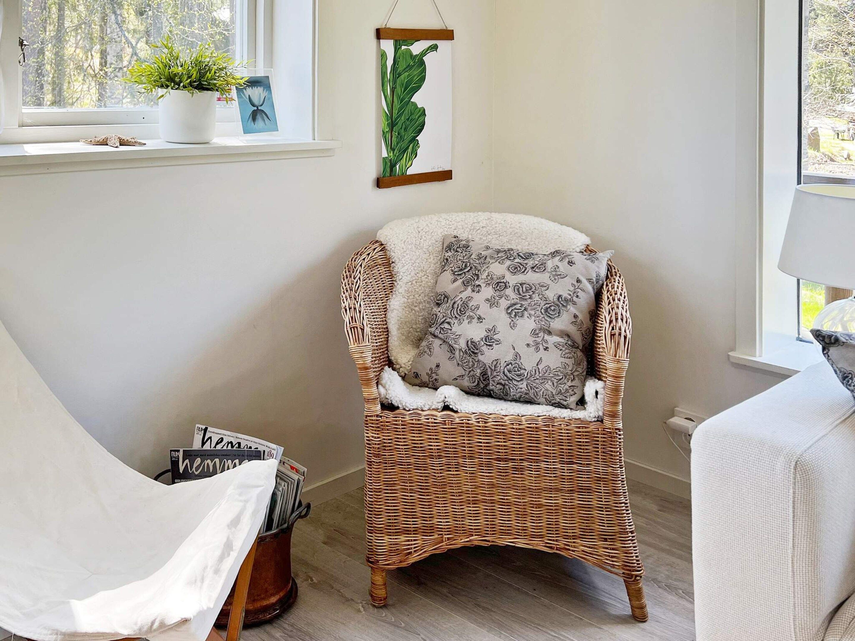 A wicker chair with a floral pillow, a potted plant, and a framed botanical print on a wall.
