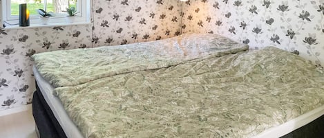 A bedroom with a bed, floral wallpaper, a window with plants, and a wall clock.