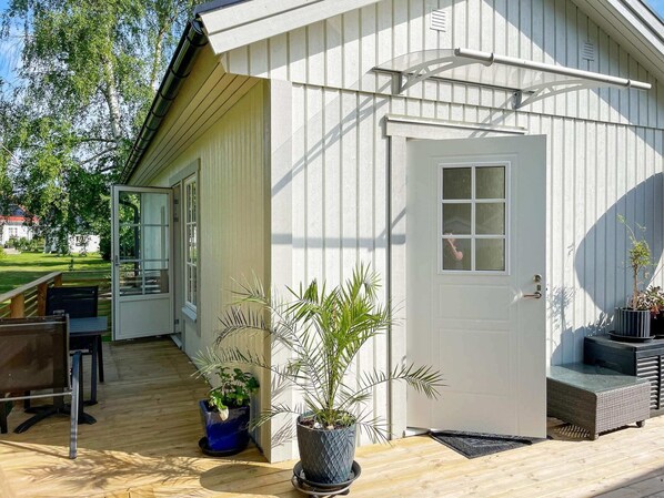 A white building with a door and windows, a wooden deck with a chair and potted plants, and a view of a house in the background. - 4 Star Holiday Home in Solvesborg (Sölvesborg)