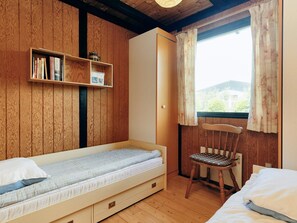 Une chambre avec un lit simple, une chaise en bois, une bibliothèque et une fenêtre avec des rideaux.