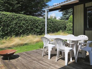 Un patio exterior con terraza de madera, mesa y sillas de plástico blanco, y una parrilla.