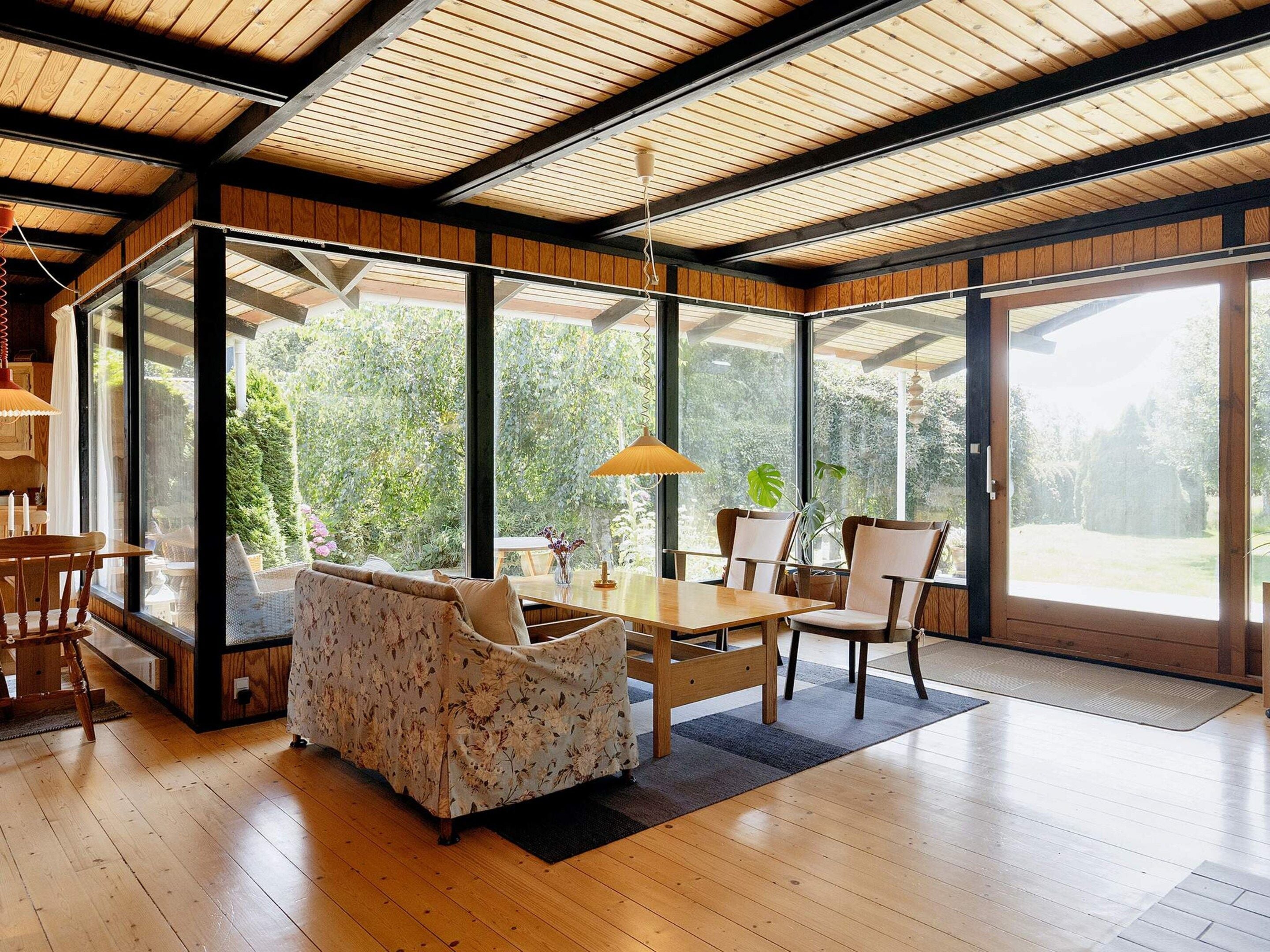 Une pièce spacieuse dotée d’un plancher en bois, d’une table à manger avec des chaises, d’un canapé et de grandes fenêtres donnant sur un paysage verdoyant.