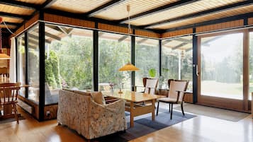 Um quarto amplo com piso de madeira, uma mesa de jantar com cadeiras, um sofá e janelas grandes que oferecem vista para o verde lá fora.