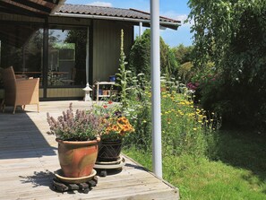 En træterrasse med plantekrukker, en fletstol og en bygning med tegltag i baggrunden.