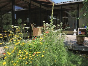 A garden with various flowers and plants, a wooden deck, and a modern building with large windows. - 7 Person Holiday Home in Karrebaeksminde (Karrebæksminde)