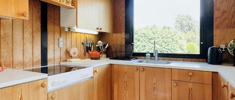 Una cocina con armarios de madera, una encimera blanca, un ventanal con vistas a zonas verdes y una lámpara colgante.