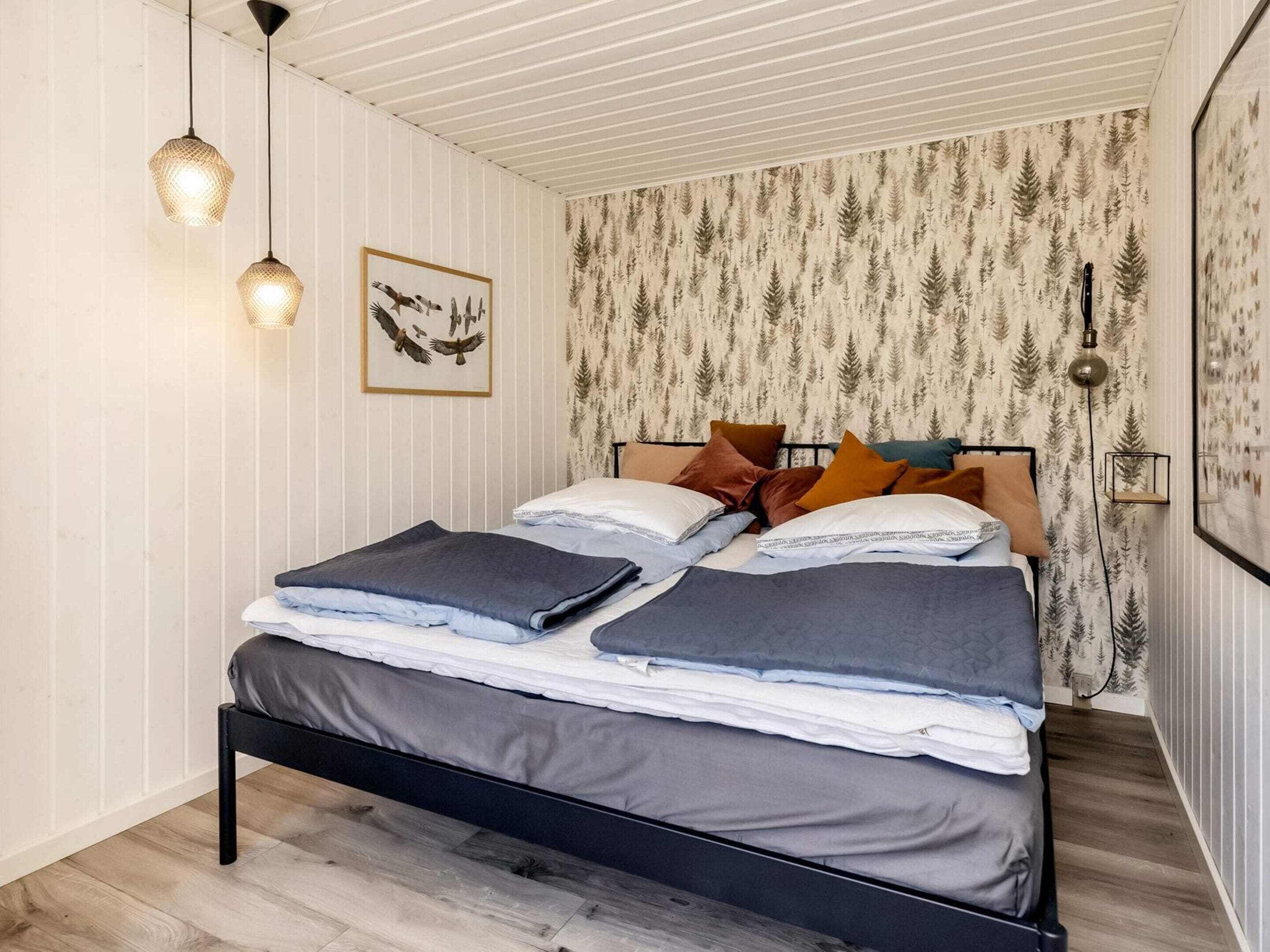 A bedroom with a bed, two hanging pendant lights, a framed picture of birds, and wallpaper with a tree pattern.