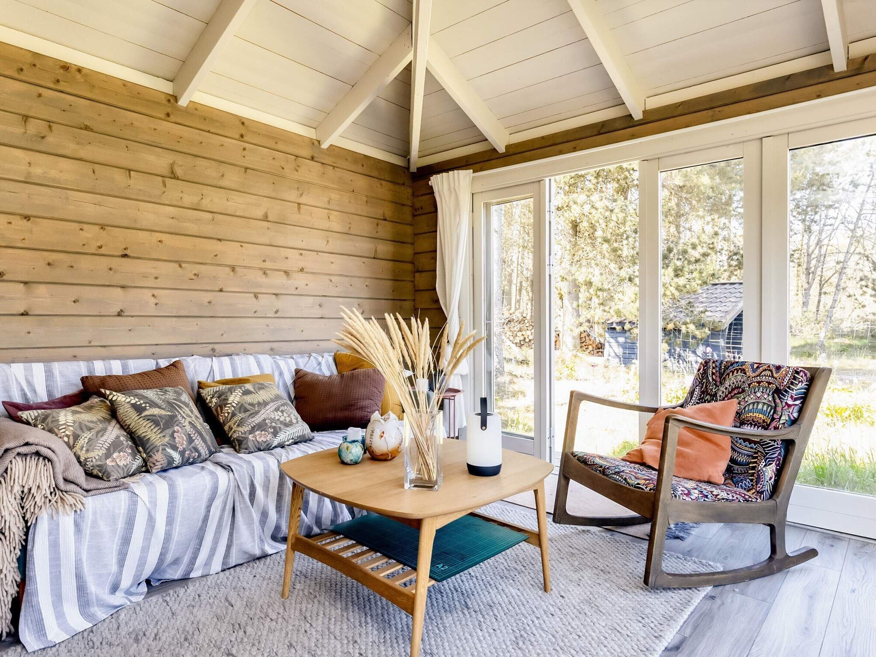 A cozy room with a wooden ceiling, a sofa with patterned cushions, a rocking chair, and a table with decorative items.