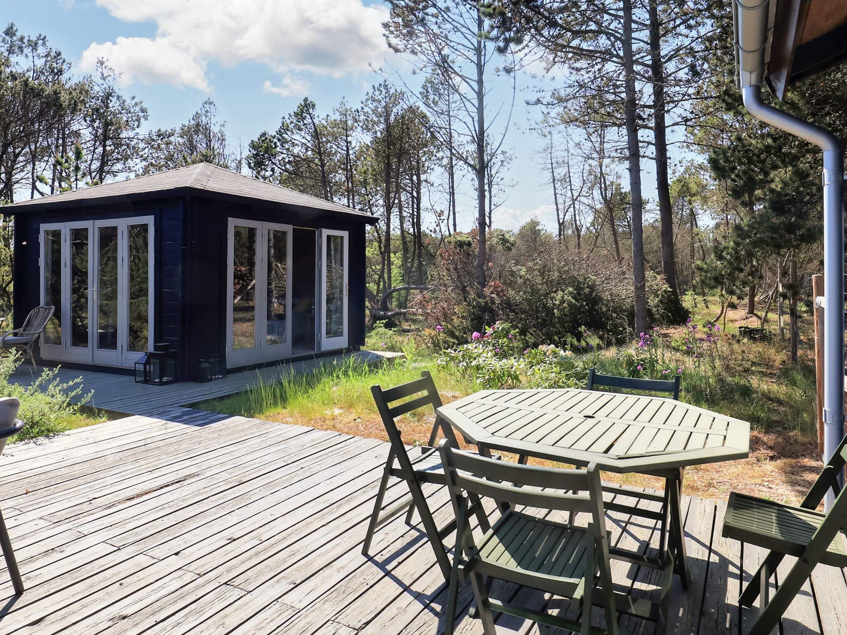 A wooden deck with a table and chairs, a small cabin with glass doors, and a forested background.