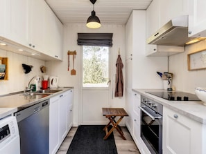 A compact kitchen with white cabinetry, a built-in dishwasher, an oven, and a window with a view of trees. - 6 Person Holiday Home in Albaek (Aalbaek)