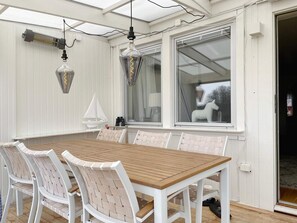 Une terrasse couverte avec une table en bois et des chaises blanches, une grande fenêtre et des lanternes suspendues.