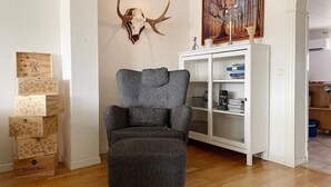 Une pièce comprenant un fauteuil gris, un crâne de cerf accroché au mur, une bibliothèque et un tableau encadré.