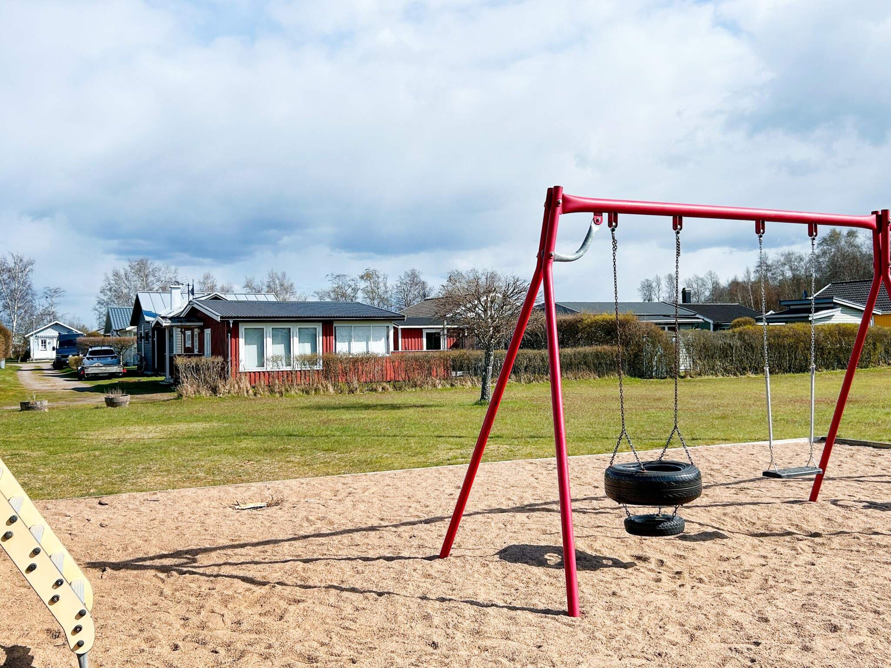 A playground with a red swing set and tire swings, surrounded by a sandy area with houses in the background.