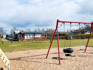 A playground with a red swing set and tire swings, surrounded by a sandy area with houses in the background. - 6 Person Holiday Home in Laholm (Skummesloevsstrand)