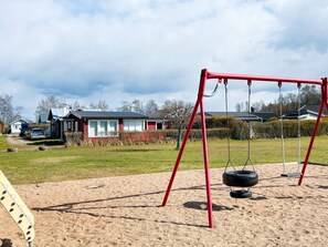 A playground with a red swing set and tire swings, surrounded by a sandy area with houses in the background. - 6 Person Holiday Home in Laholm (Skummesloevsstrand)
