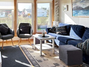 Een woonkamer met een blauwe bank, twee fauteuils, een salontafel en uitzicht op een woonwijk door grote ramen.