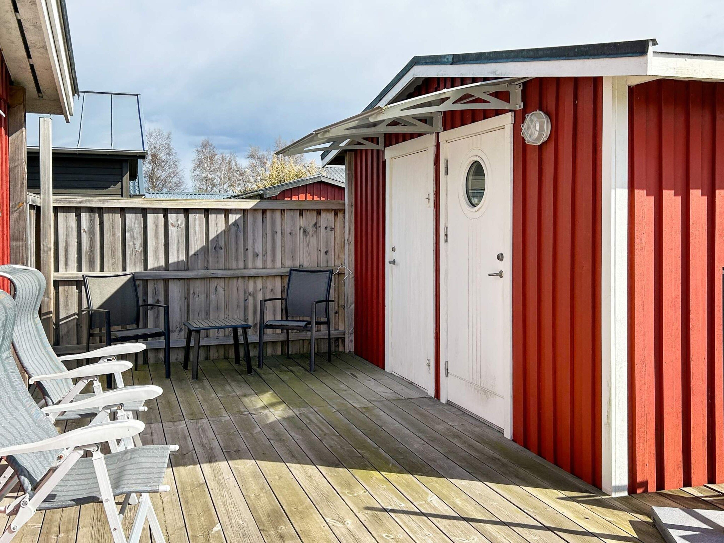 Una terraza de madera con mobiliario exterior y un edificio rojo con puerta y ventana.