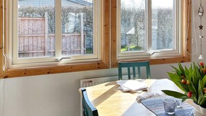 A room with a table and chairs, a window with a view of a wooden fence and trees, and a vase with flowers. - 6 Person Holiday Home in Laholm (Skummesloevsstrand)
