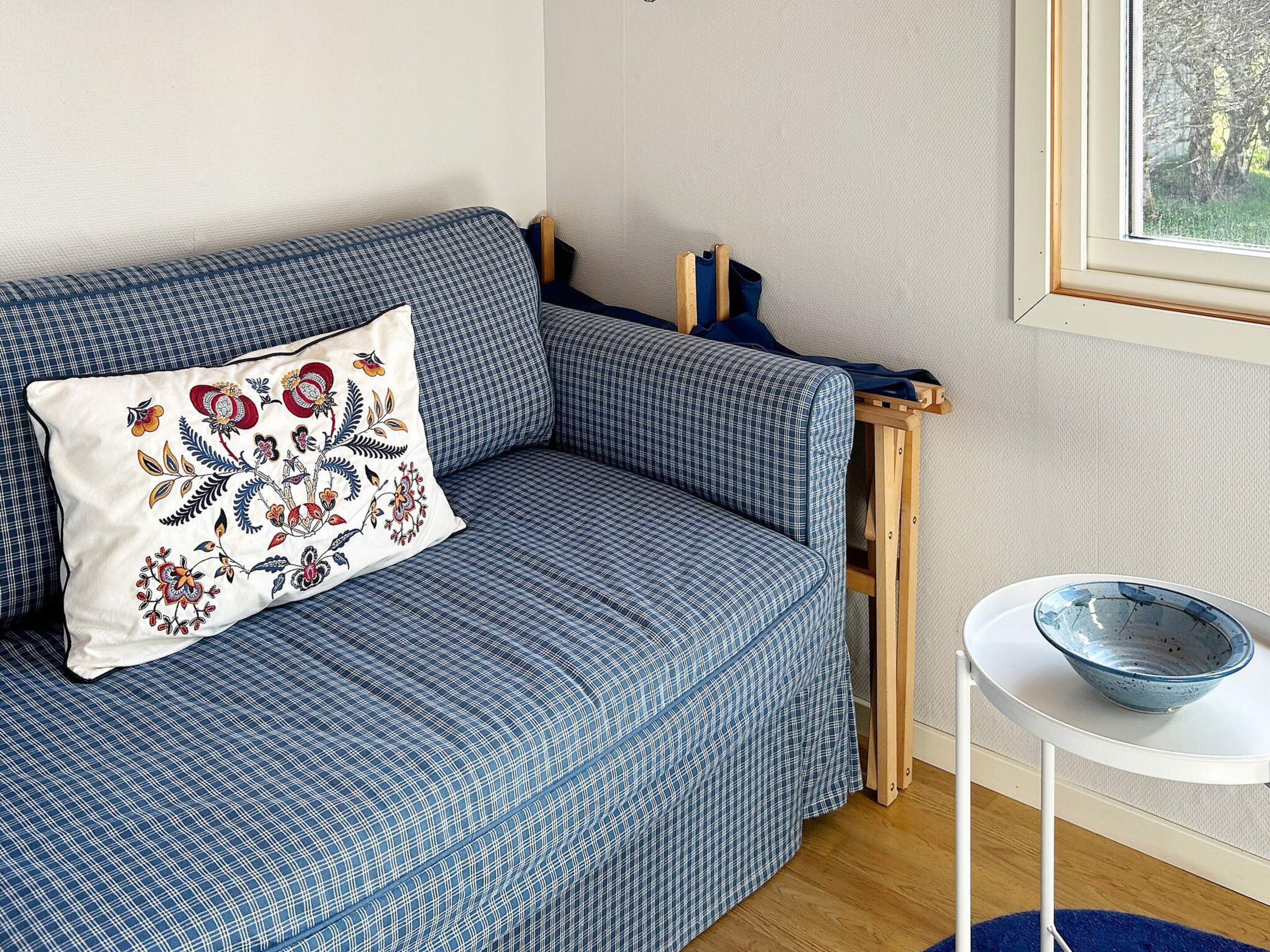 A blue sofa with a decorative pillow, a small white table with a bowl, and a window with a view of trees.