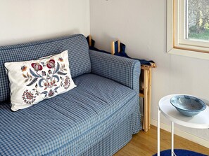 A blue sofa with a decorative pillow, a small white table with a bowl, and a window with a view of trees. - 6 Person Holiday Home in Laholm (Skummesloevsstrand)