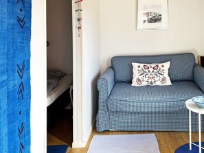 Quarto com sofá xadrez, uma mesinha branca, quadro pendurado na parede e cortina azul.
