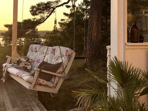 A wooden swing chair with floral upholstery, a wooden deck, and a view of trees and a building in the background. - 4 Star Holiday Home in Uddevalla (Uddevalla)
