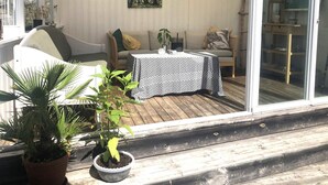 Une terrasse ensoleillée avec une plateforme en bois, une table recouverte d’une nappe à motifs, un canapé avec des coussins et une plante en pot.