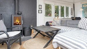 A cozy room with a wood-burning stove, a sofa, a dining table, and a window with a view of greenery. - 5 Person Holiday Home in Thyholm (Thyholm)