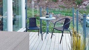 En terrasse med to stole og et lille bord, omgivet af glasgelændere og udsigt til grønne omgivelser.