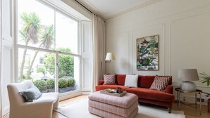 A living room with a red sofa, a beige armchair, a striped ottoman, and a painting of a traditional building. - Pembridge Villas VI by onefinestay (London)