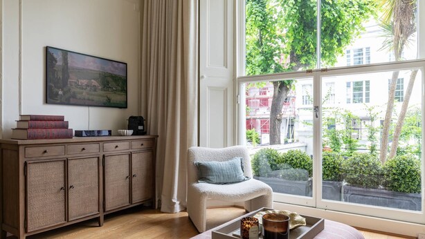 A cozy room with a large window, a wooden cabinet, a chair, and a tray with candles. - Pembridge Villas VI by onefinestay (London)