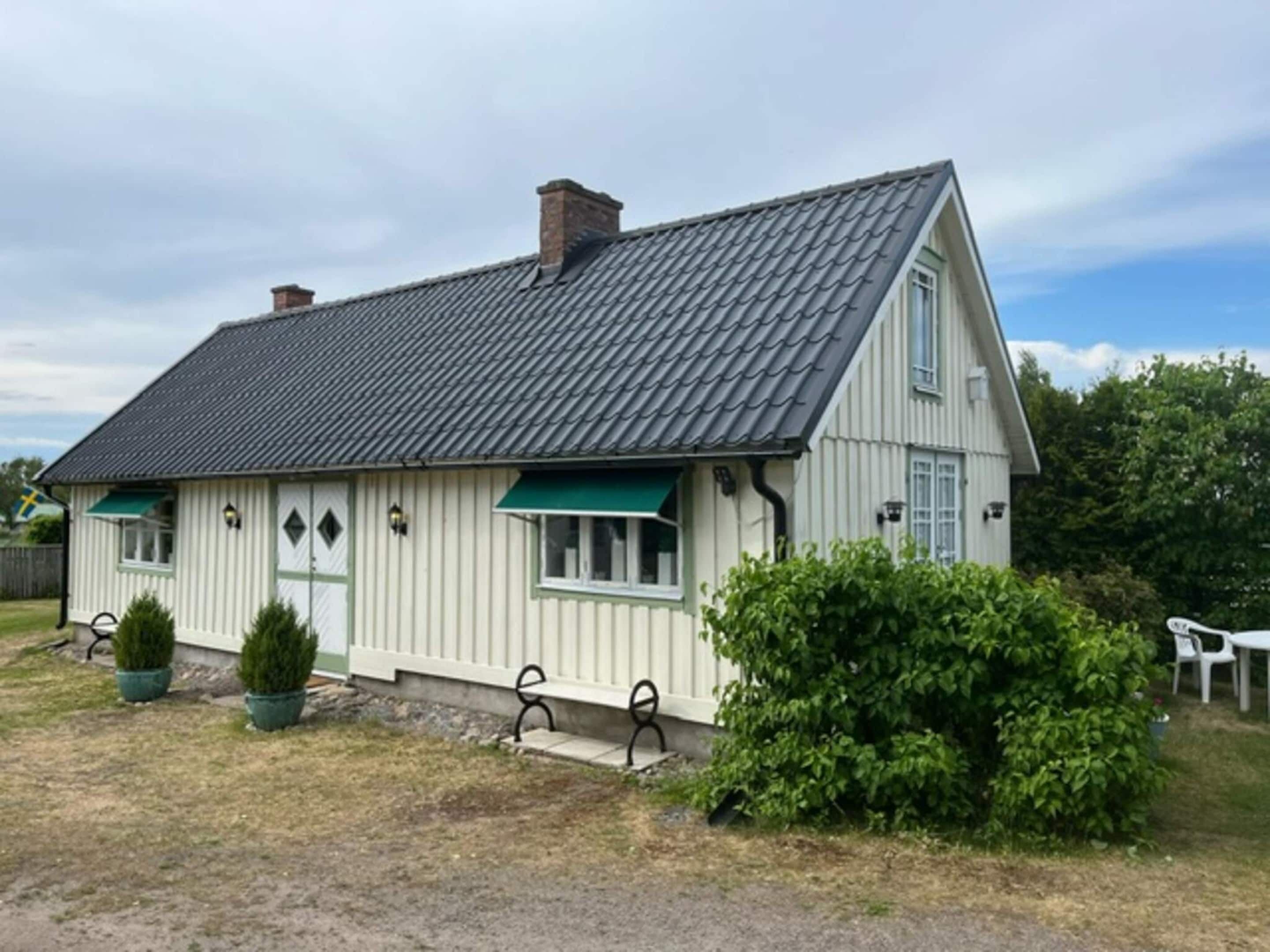 A single-story house with a sloped roof, a front porch with a bench, and a small garden area with potted plants.