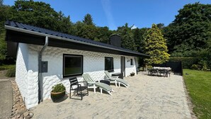 Une maison moderne avec une façade en pierre, un patio agrémenté de mobilier d’extérieur et un grand arbre en arrière-plan.