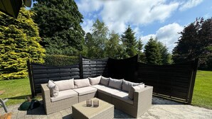 Un canapé d’angle en osier avec des coussins, une table basse en fibres tressées et une chaise en métal sur une terrasse en pierre.