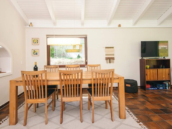 A dining area with a wooden table and chairs, a window with blinds, a wall-mounted TV, and a bookshelf with books and a bag.