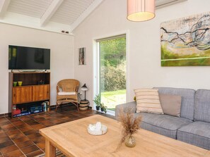Una sala de estar con un sofá, una mesa de centro de madera, un televisor sobre un mueble, una silla de mimbre y un cuadro en la pared.