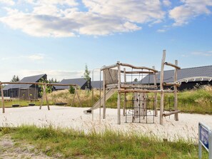 Un terrain de jeux avec des balançoires et un toboggan, entouré de sable et d’herbe, avec des bâtiments en arrière-plan.