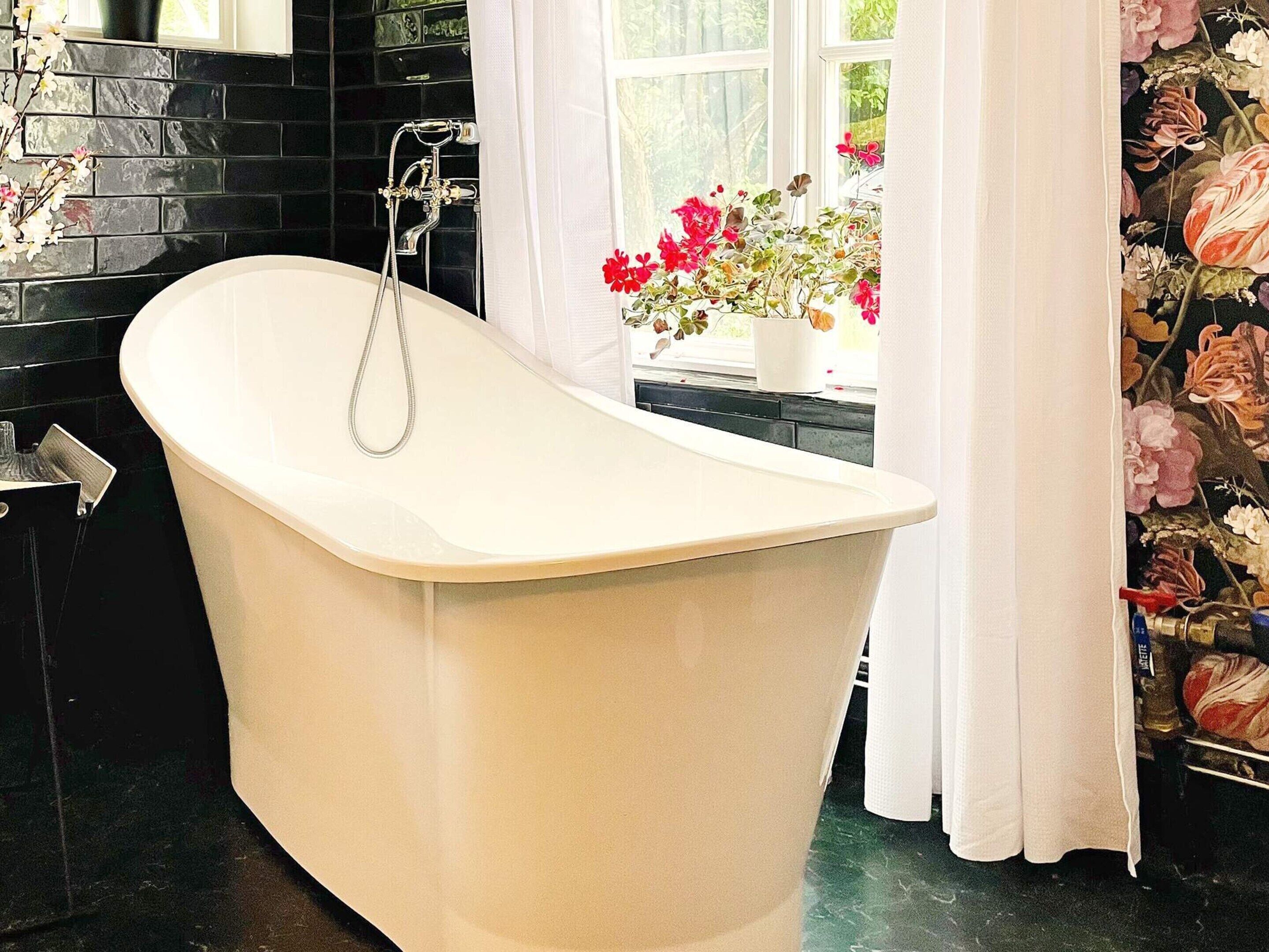 A freestanding bathtub in a bathroom with a black tiled wall and a window with a view of flowers.