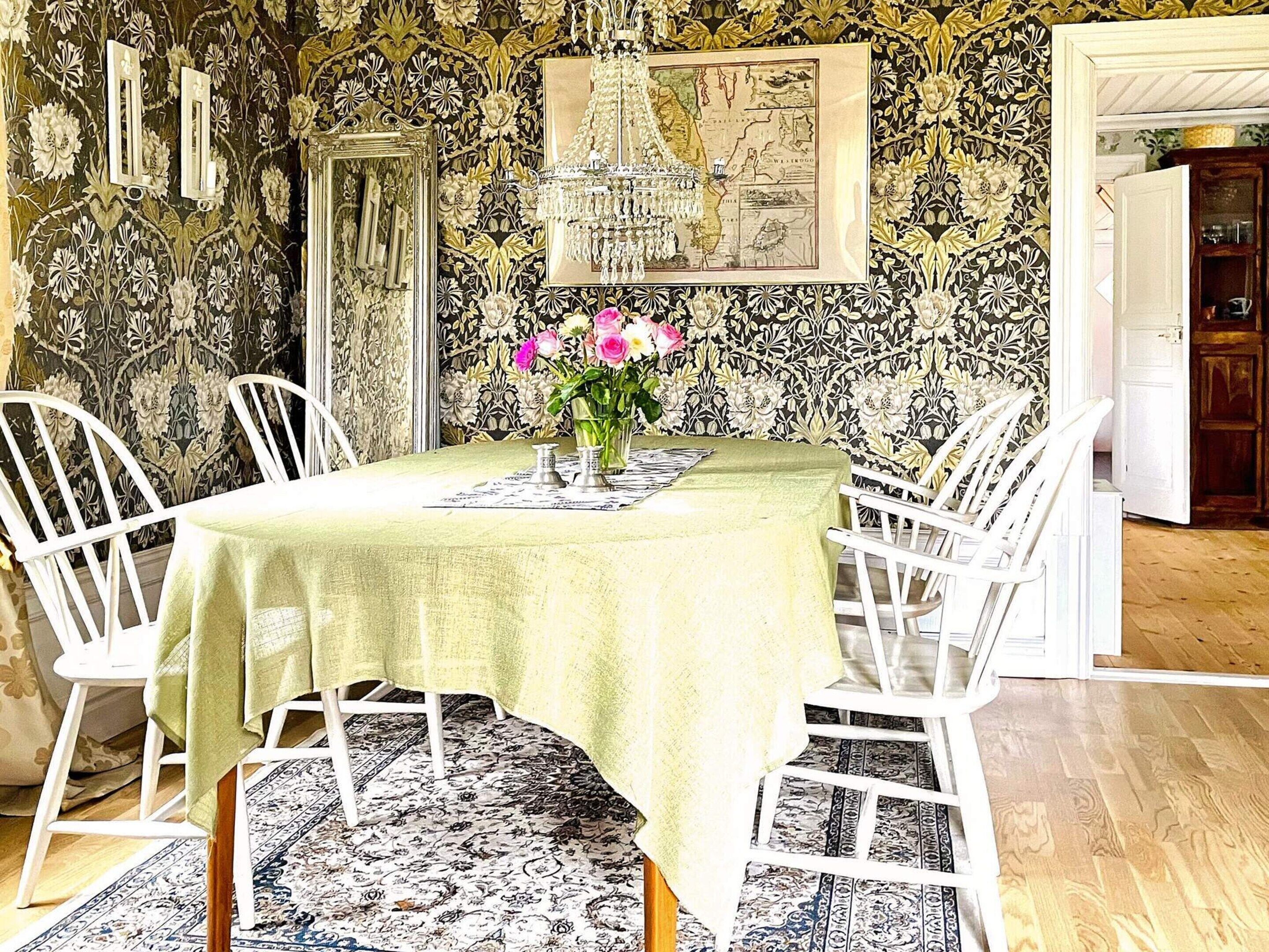 A dining area with a table set for four, surrounded by white chairs, a patterned rug, and a floral wallpaper.