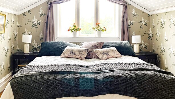 Een slaapkamer met een groot bed, twee nachtkastjes met lampen, een raam met gordijnen en bloemenbehang.