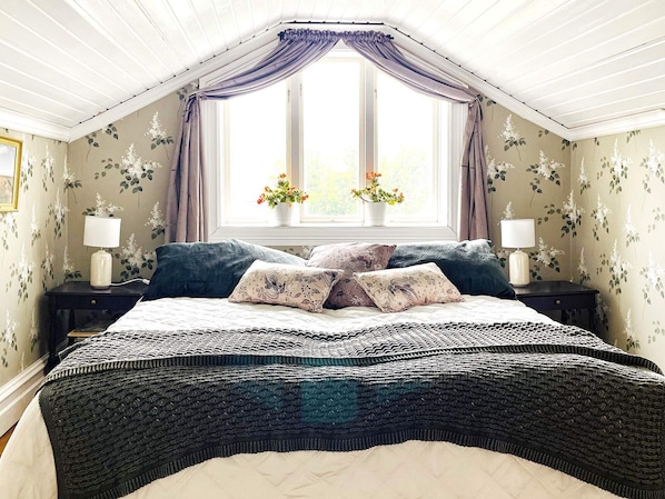 Une chambre à coucher avec un grand lit, deux tables de chevet avec des lampes, une fenêtre avec des rideaux et un papier peint à motifs floraux.