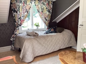A bedroom with a bed, floral curtains, a window with a plant, a wooden floor, and a door.