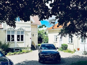 Une rue résidentielle où est garée une voiture noire devant une maison à la façade blanche et au toit recouvert de tuiles rouges.