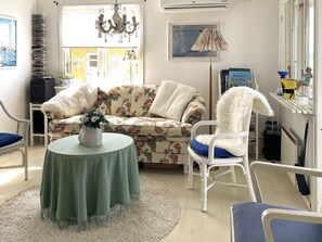 Un salon avec un canapé à motifs floraux, un fauteuil blanc, une table ronde ornée d’un vase, un lustre et un tableau encadré accroché au mur.