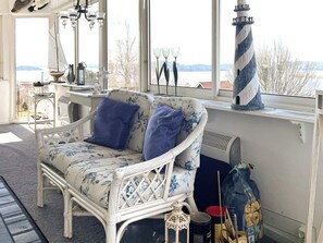A wicker sofa with floral upholstery and blue cushions, a white lamp, a lighthouse model, and a view of a body of water through large windows.